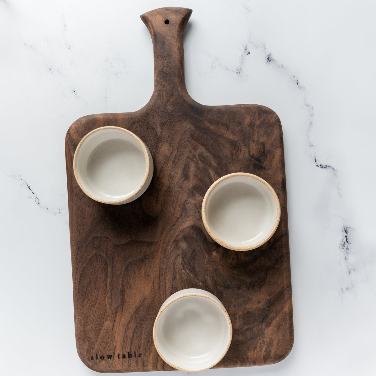 A paddle style walnut serving board styled with three handmade stoneware ramekins, placed on a bright white marble countertop.