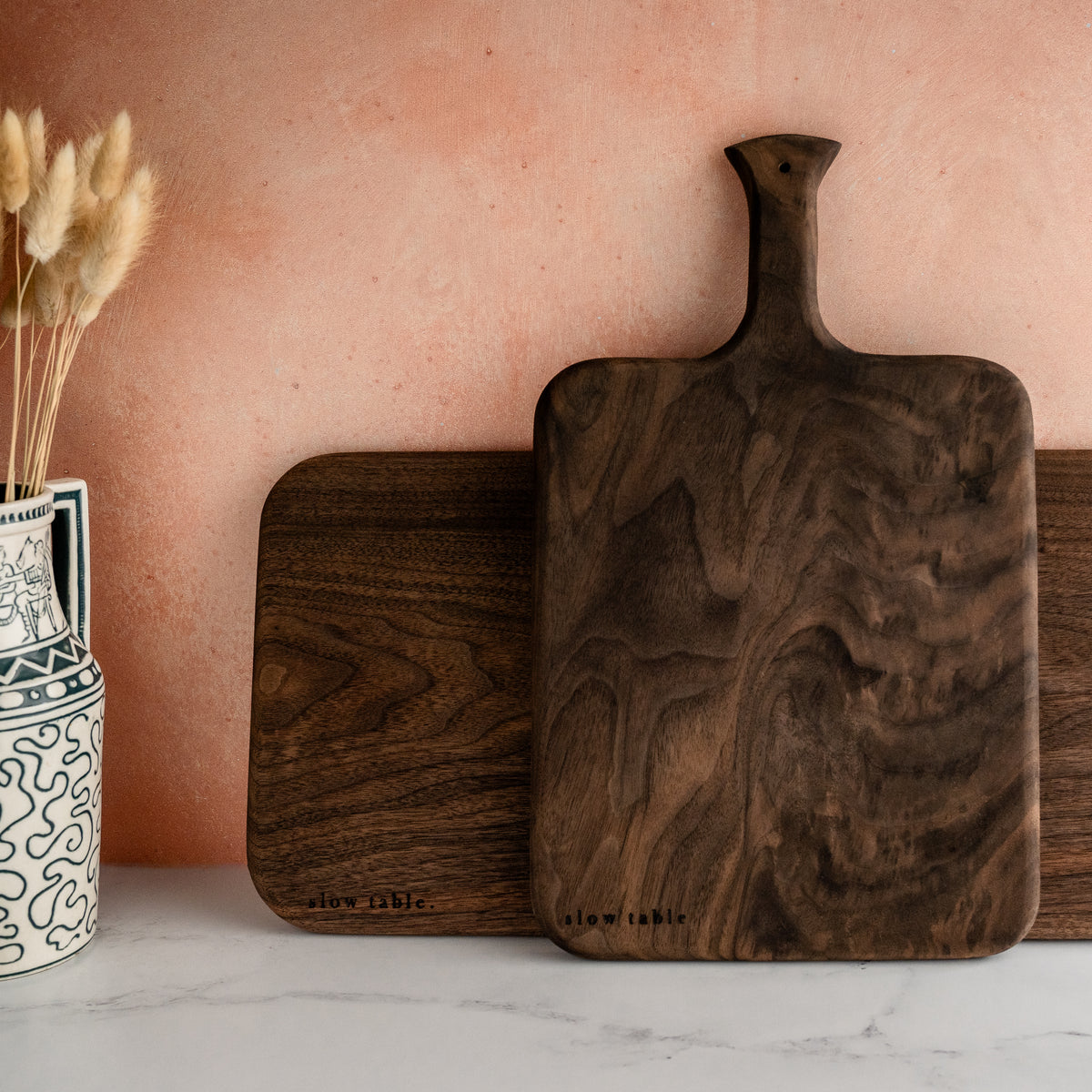 image of two sizes of solid walnut wood boards, propped against a kitchen backsplash on a marble countertop. Both boards have a beautiful fine and unique wood grain.
