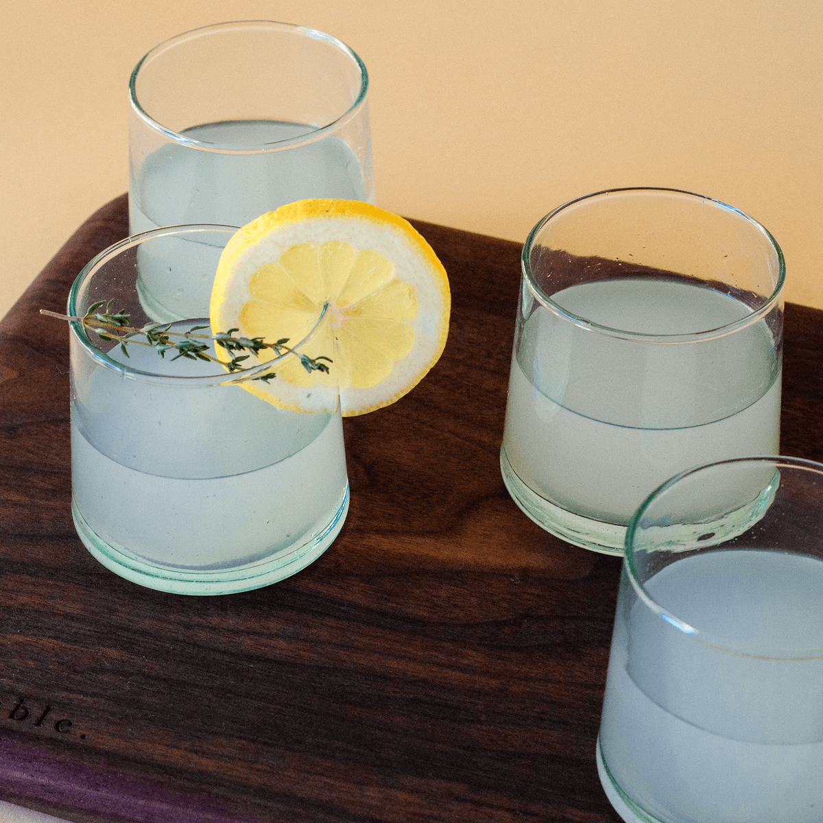 small handblown drinking glasses, filled with a cocktailed beverage and garnished with lemon slice and thyme, sitting on a solid walnut wood serving board.