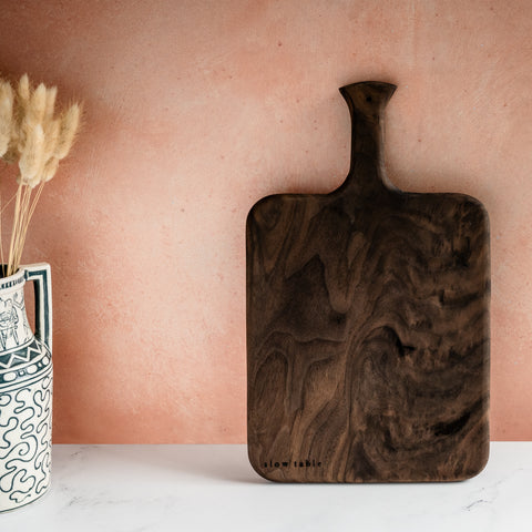 A beautiful paddle-style wood serving/cutting board made of solid walnut, with a beautiful grain, propped against a kitchen backsplash and resting on a marble countertop. Pictured with dried grasses in a vase.