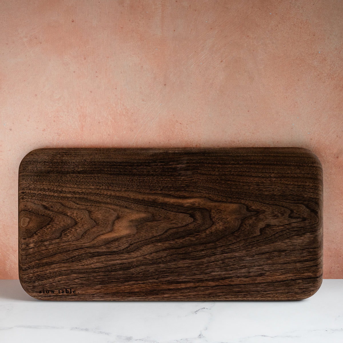Rectangular, solid walnut wood serving/cutting board, with a fine wood grain, propped against a kitchen backsplash and resting on a marble countertop.